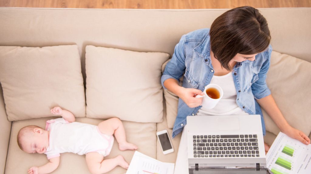 Woman sitting on a couch while working on a notebook, drinking tea, and taking care of a baby
