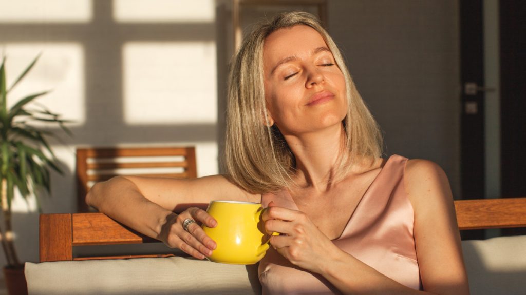 Happy woman sitting at home, relaxing, and enjoying the sun and a cup of coffee or tea
