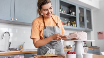 culinary, baking and cooking food concept - happy smiling young woman making layer cake and spreading topping cream on kitchen at home