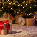 A Christmas tree with lights. A few presents on a carpeted floor protector