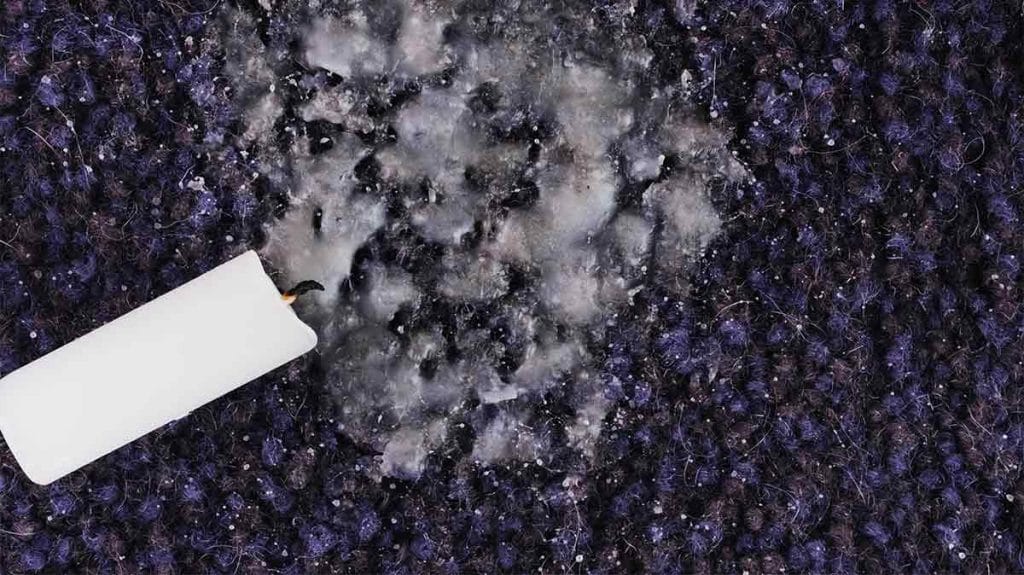 A white candle which has fallen and spilled wax on a purple carpet.