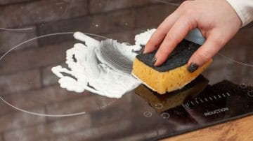A hand cleaning an induction stove top with a yellow sponge.